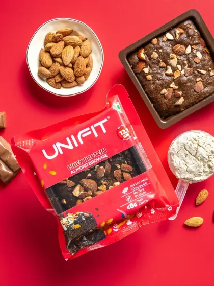 A packet of almond-flavored whey protein brownie, shot against a bold red background with almonds and chocolate.