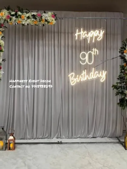 For a more elegant and sophisticated theme, I can create backdrops using drapes and flowers. This setup for a 90th birthday features a white curtain, floral arrangements, lanterns, and a custom neon sign.