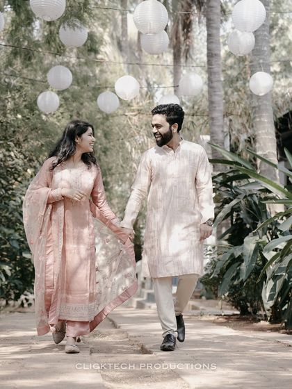 A couple in traditional pastel outfits walks hand-in-hand down a path decorated with hanging paper lanterns.