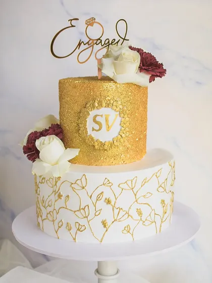A glamorous two-tiered engagement cake featuring a brilliant gold sequin top tier and a hand-painted gold floral pattern on the bottom. The custom "SV" monogram adds a personal touch.
