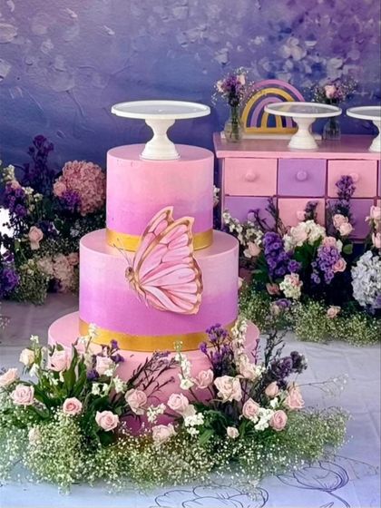 A close-up of the beautiful two-tiered cake, decorated with a delicate butterfly. The cake is surrounded by fresh flowers, perfectly matching the party's pink and purple floral theme.