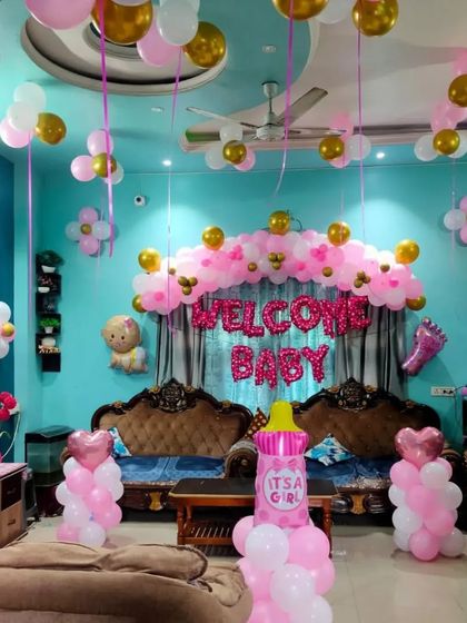A full room decoration to welcome a baby girl, with pink and gold balloons, "Welcome Baby" foil letters, and balloon columns.