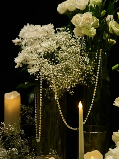 A close-up showing the textures of baby's breath, white roses, and a string of pearls, all illuminated by candlelight.