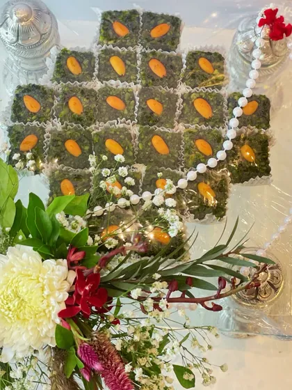 A traditional mithai platter featuring delicious green sweets topped with almonds, arranged on a clear tray with silver accents and a fresh floral bouquet.
