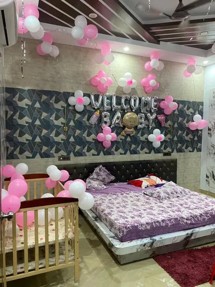 A full bedroom decoration to welcome a baby girl. The setup includes "Welcome Baby" foil balloons over the bed, a decorated crib, and pink and white balloon flowers on the walls and ceiling.