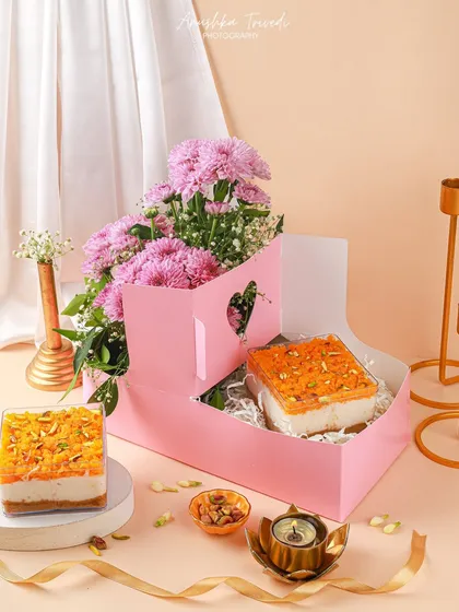 A beautiful pink hamper box designed for gifting, containing two tubs of Indian fusion dessert. The composition with flowers and candles creates a soft, celebratory mood.