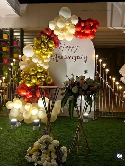 An outdoor anniversary decoration featuring a white arched backdrop with custom text. The backdrop is framed by a balloon garland in red, gold, and cream, perfect for a garden or terrace party.
