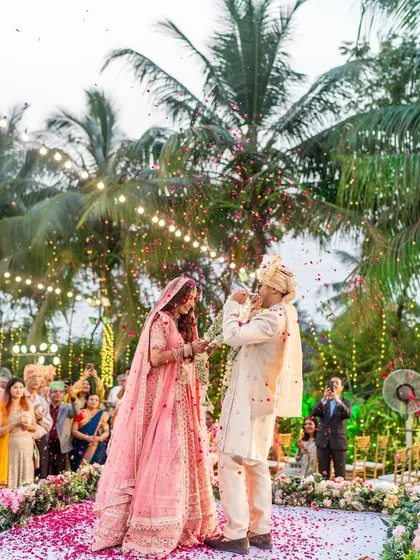 A beautiful outdoor varmala ceremony surrounded by palm trees. The playful moment of the groom bowing his head for the garland exchange is full of fun and tradition.
