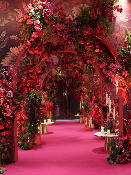 A view down the dramatic red floral entrance tunnel. The path is lined with candles and golden vases, enhancing the warmth and luxury of the design. The floral wallpapered walls add another layer of texture to this immersive experience.