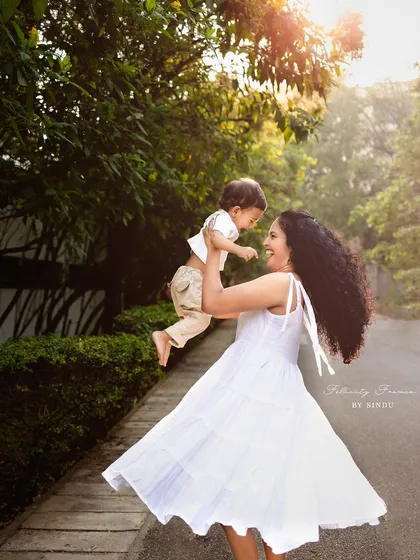 A mother in a white dress joyfully lifts her child into the air during a sunlit outdoor session. This image is full of movement and happiness, capturing a perfect moment of motherly love.