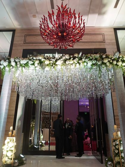 A glamorous entrance detail featuring a ceiling of hanging silver crystals and white florals, set under the venue's dramatic red chandeliers. This design is all about shimmer and sophistication.
