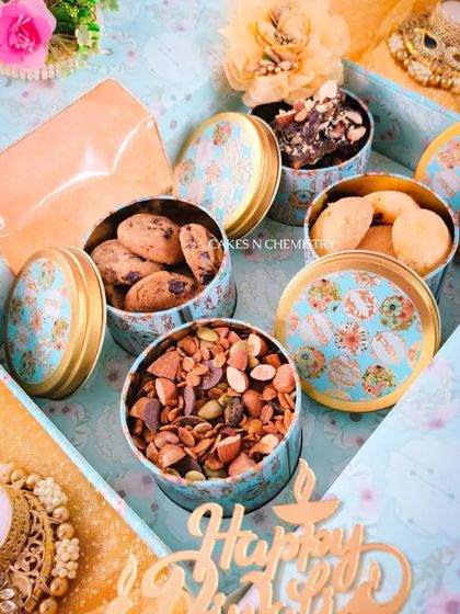 This beautiful Diwali hamper features an assortment of treats in elegant tin boxes, including chocolate chunk cookies and a premium trail mix. The box is accompanied by a decorative diya, making it a complete festive gift.