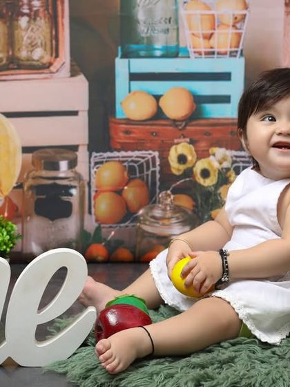 A fruity and fun first birthday. This little one is celebrating her milestone surrounded by fresh fruits and a rustic, market-themed backdrop, showing off her happy smile.