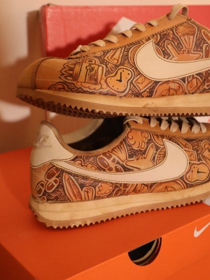 A detailed close-up of the travel doodle on the Nike Cortez. The coffee-dyed surface enhances the hand-drawn quality of the art, making it look like a well-loved sketchbook.