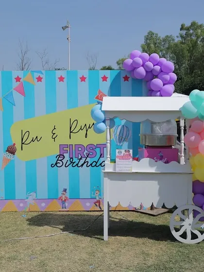 An outdoor candy floss station designed for a twins' first birthday carnival. The setup includes a custom backdrop with their names, a classic white cart, and playful pastel balloon garlands.