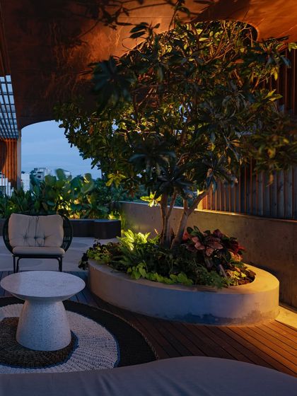 A cozy seating area on a rooftop at night, with a tree in a large planter and surrounding greenery softly lit. The design creates an intimate and peaceful corner to enjoy the evening air.