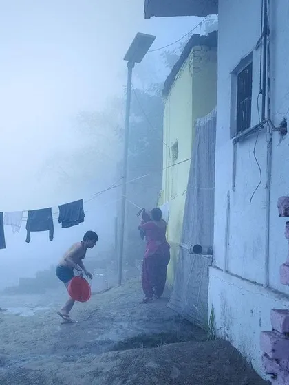 Life unfolds on a foggy morning at the ghat, with laundry hanging to dry and people starting their day, all enveloped in a thick, blue-toned mist.