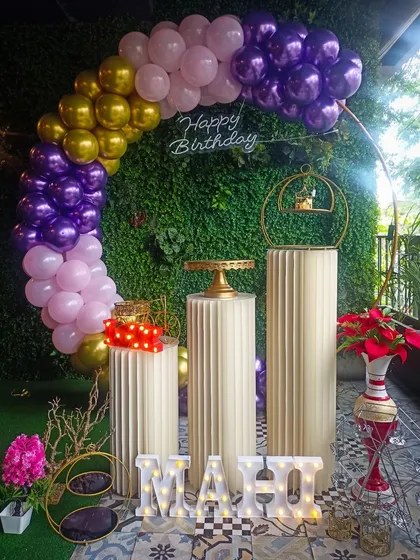 This vibrant balloon arch uses a rich color palette of purple, pink, and gold against a lush green wall. The setup includes classic pedestals for the cake, a neon sign, and light-up marquee letters for the name 'MAHI'.
