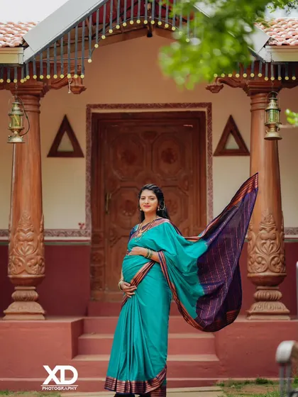 A stunning solo portrait of the mother-to-be. The flowing teal saree against the rustic backdrop of a traditional home creates a picture of pure elegance and grace.