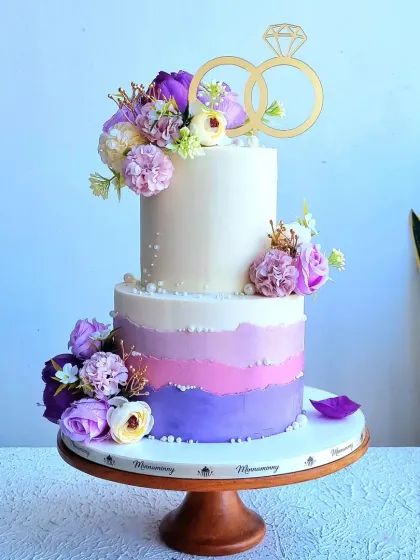An elegant two-tier cake ideal for a wedding or engagement celebration. The bottom tier features a beautiful purple ombre effect, and the cake is decorated with artificial flowers and a classic ring topper, offering a sophisticated and modern design.