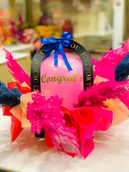 A vibrant congratulations gift hamper. This arrangement features a large pink balloon and is decorated with brightly colored dried flowers and ribbons, all in a stylish black carrier.