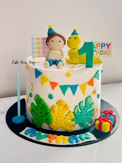 A whimsical first birthday cake with a cute fondant boy and his dinosaur friend, decorated with tropical leaves and colorful bunting.