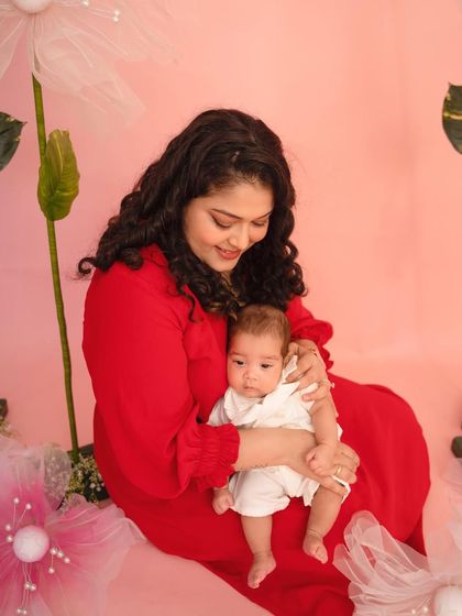 A mother's gentle hold and loving smile are the focus of this beautiful portrait, set against a soft pink backdrop with floral accents.