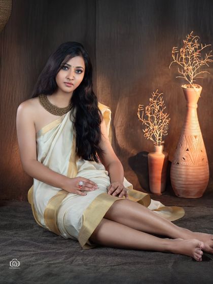 A seated portrait that captures the simple elegance of the Kerala saree. The warm, earthy tones of the studio backdrop complement the gold border of the saree, creating a serene and beautiful image.