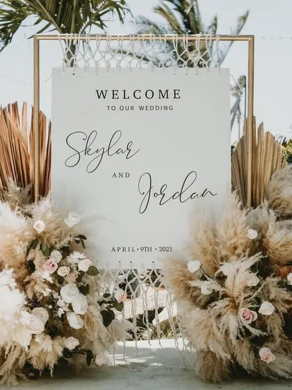 This welcome sign for a beach wedding features a modern design with pampas grass and dried palm leaves. The bohemian-chic aesthetic perfectly complements the natural, sandy surroundings.