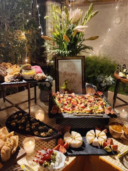 An evening view of a beautiful grazing table set up in a garden for a Diwali party. The fairy lights and candles add a magical touch to the spread of cheeses, breads, dips, and appetizers.
