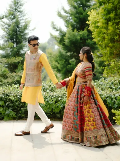 A beautiful portrait of the couple during their Haldi, holding hands and walking through a lush green path. The vibrant colors of their outfits pop against the natural backdrop.