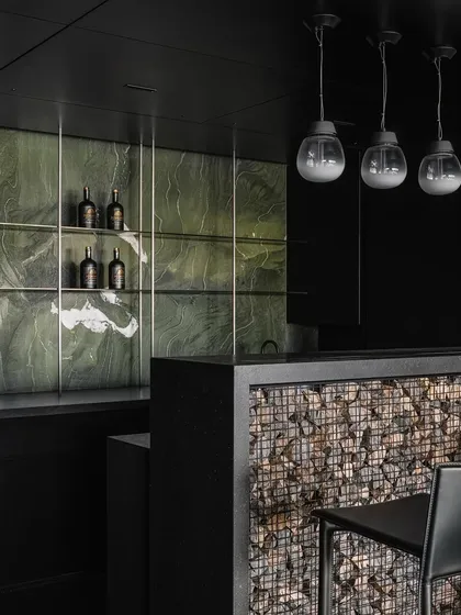 We believe in the honest expression of materials. For this bar area, we conceptualized a Gabion wall, trapping randomly sized stones in a metal wire mesh to create a raw, textural fascia. This Brutalist element is contrasted with a sleek, dark green marble backsplash and minimalist pendant lighting, forming part of a 'black scoop' that unifies the entrance, bar, and kitchen.