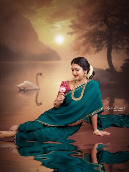 A fine art composition with a painterly feel. The mother-to-be, in her green saree, interacts with a flower by a dreamy, swan-filled lake at sunset.