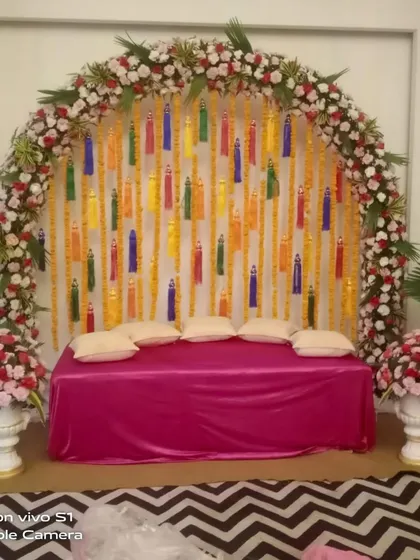 An indoor floral arch with colorful tassels hanging against a white backdrop. This is a great way to add a pop of color to your Mehndi or Haldi decor.