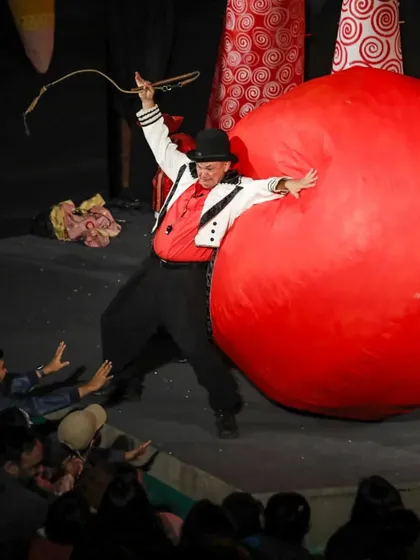 The ringmaster playfully battles a giant red inflatable ball, a moment of pure circus clowning that has the audience roaring with laughter.