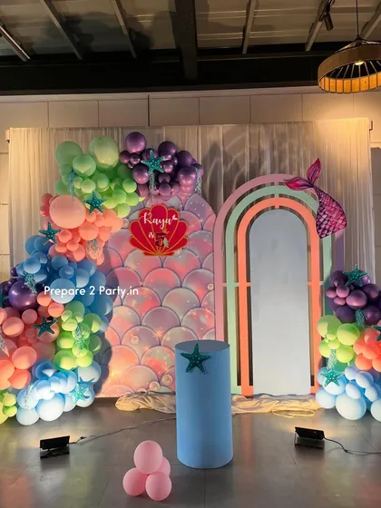 Dive into an under-the-sea adventure with this complete mermaid theme decoration. It features a shimmering scale backdrop, a pastel rainbow arch, and a colorful balloon garland with starfish and mermaid tail details.