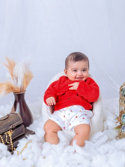 A happy baby in a red sweater, sitting in a tiny armchair on a bed of clouds. The simple props like the treasure chest and birdcage add a touch of vintage charm.