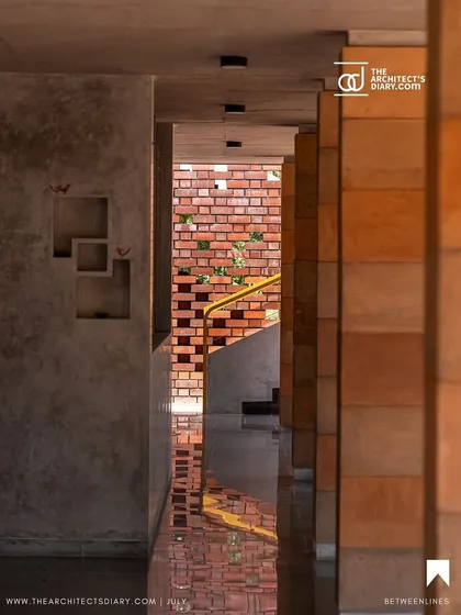 A view through the colonnade of CSEB pillars at "Aamra." The polished floor reflects the exposed brick of the staircase wall, showcasing the raw, honest material palette of cement, earth, and stone.