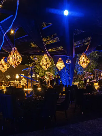 A wider view of the outdoor seating area, showing the interplay of the dark draped ceiling, warm chandeliers, and cool blue accent lighting.