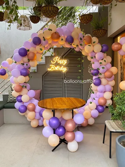 This shows my work in a cafe setting, creating a beautiful birthday photo corner. The design uses a soft pastel palette of purple, peach, and rose gold balloons in a circular arch around a table, complete with a neon sign.
