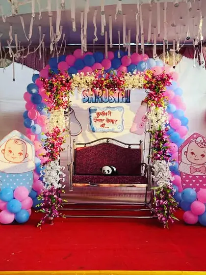 A large outdoor naming ceremony setup. The stage has a grand entrance arch made of balloons and flowers, leading to the decorated jhula.