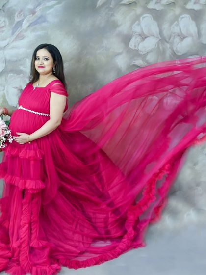 A dynamic studio shot featuring a mom-to-be in a vibrant pink ruffled gown. The "flying fabric" effect adds a touch of magic against the floral backdrop.