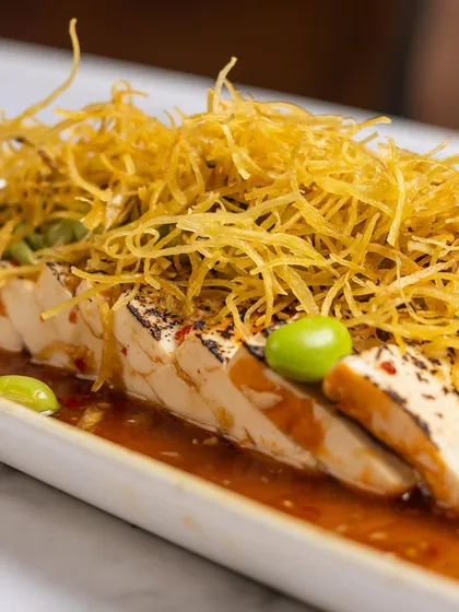 Silken tofu, but with a story of contrasts. This Tofu Carpaccio is served with a ginger ponzu dressing, avocado crema, and a pile of crispy sweet potato strings for texture. Minimal on the plate, memorable on the palate.