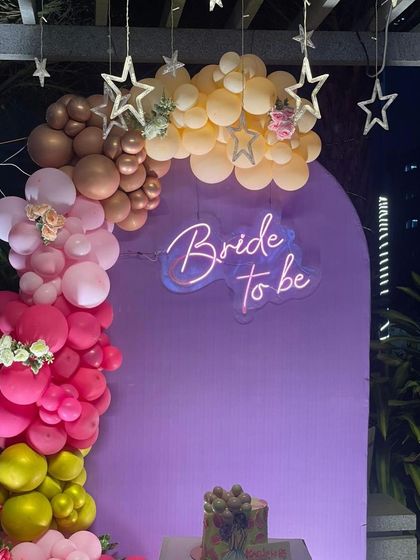 A stunning outdoor setup for a 'Bride to be' party. The purple backdrop with a neon sign is framed by a colorful organic balloon arch in shades of pink, yellow, and brown, with star lights twinkling above.
