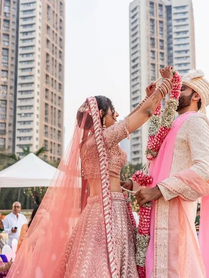 The bride reaches up to place the garland on the groom, a classic varmala moment set against a modern city backdrop.