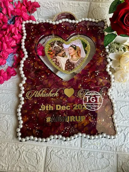 A 12x16 inch frame with a hanging heart photo cutout, set against a background of deep red preserved varmala petals. The design includes the couple's names, date, and wedding hashtag.