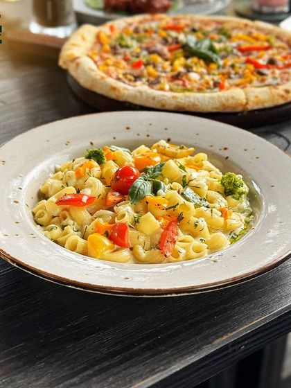 A creamy, comforting bowl of macaroni pasta with vegetables, with a fresh pizza in the background. A feast for any craving.