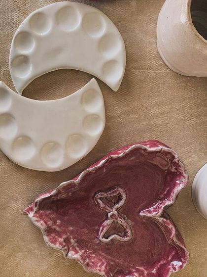 A collection of handmade dishes, including crescent-shaped paint palettes and a heart-shaped trinket dish in a deep magenta glaze. These pieces are designed to bring joy to your creative space or dressing table.