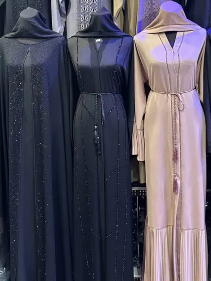 Three elegant abayas on mannequins, including a black cape style piece and two belted designs with tassel details in black and beige.