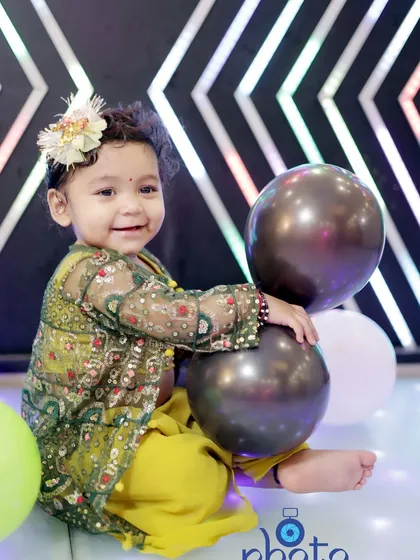 A happy baby girl in a green and yellow traditional outfit, smiling as she plays with balloons on the floor.